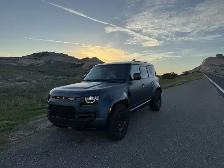 Blue Defender OCTA parked beside a rural road in a front three-quarter view at sunrise.