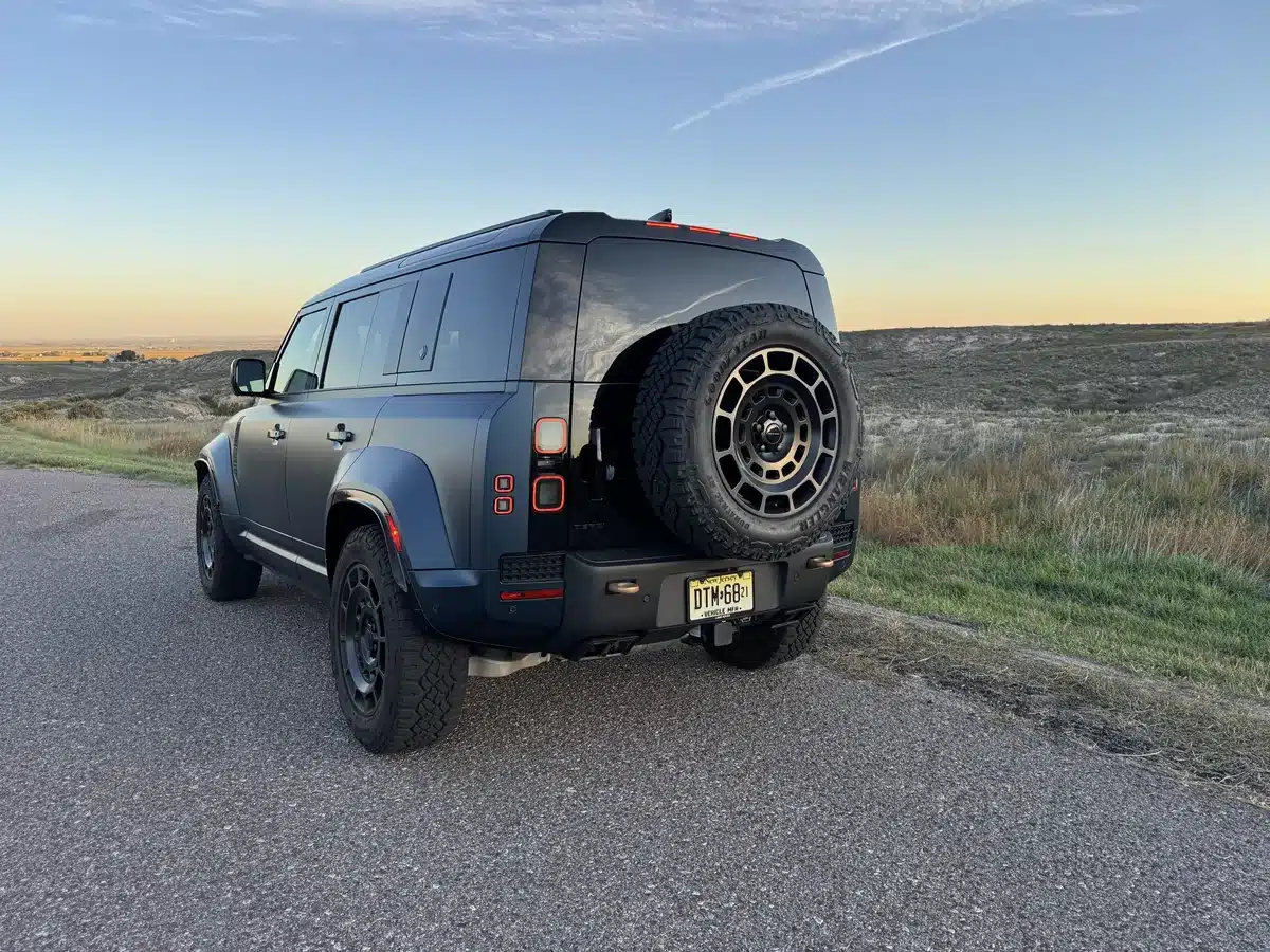 Rear three-quarter view of a blue Defender OCTA with a tailgate-mounted spare tire beside a rural road.