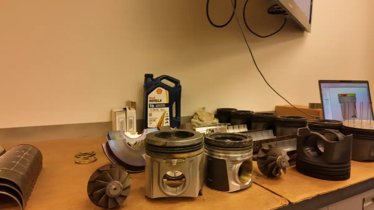 Pistons and other engine components on a Shell laboratory bench beside a computer display.